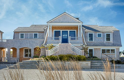 Elegant coastal home with shingle siding, blue front doors, and covered porch, featuring classic New England architecture and landscaped entrance.