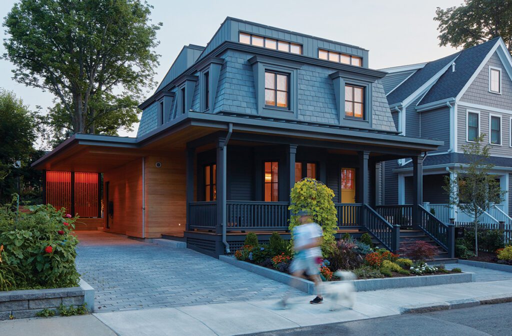 Modernized historic home with a dark gray exterior, large front porch, and wooden carport, set in a landscaped residential neighborhood, with a person walking a dog on the sidewalk.