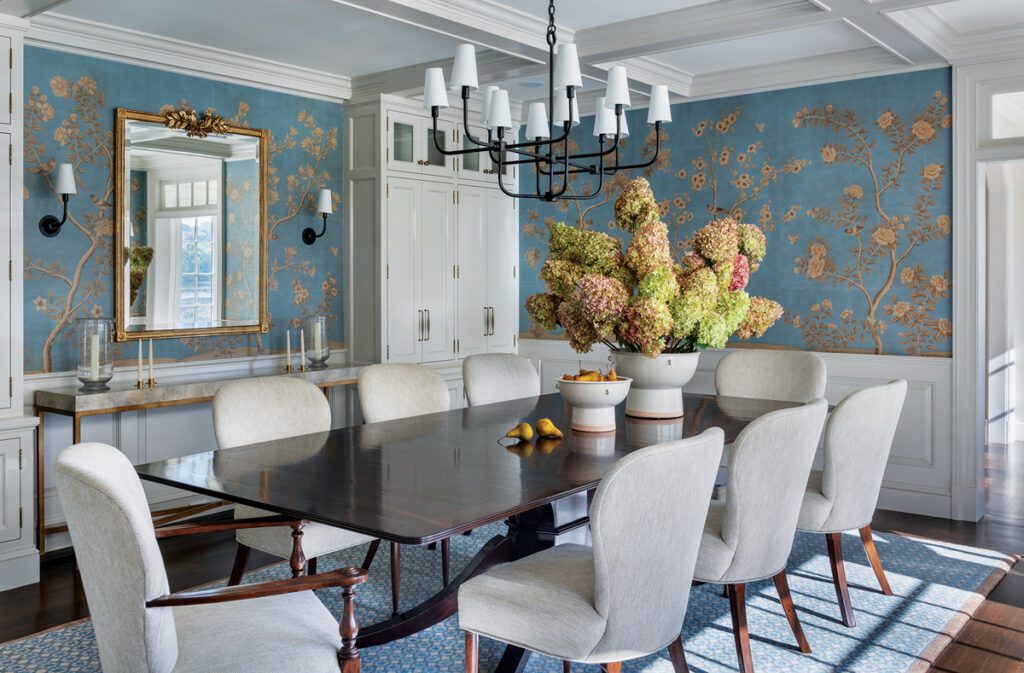 Elegant dining room with a dark wood table, light upholstered chairs, and a vibrant blue wallpaper adorned with golden floral designs. A large floral centerpiece and a gold-framed mirror add a touch of sophistication.