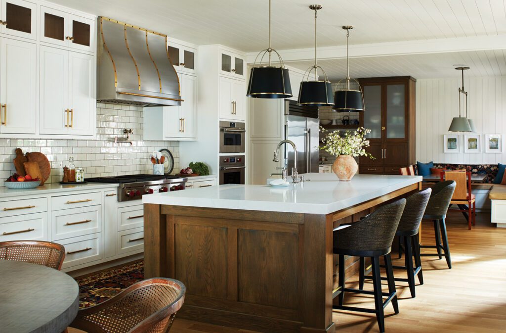 Spacious modern kitchen featuring a large wooden island with white quartz countertops, black pendant lighting, and upholstered barstools. The space includes white cabinetry with brass hardware, a custom range hood with brass detailing, glossy subway tile backsplash, and commercial-grade appliances. A cozy dining nook and built-in cabinetry complete the open and inviting design.