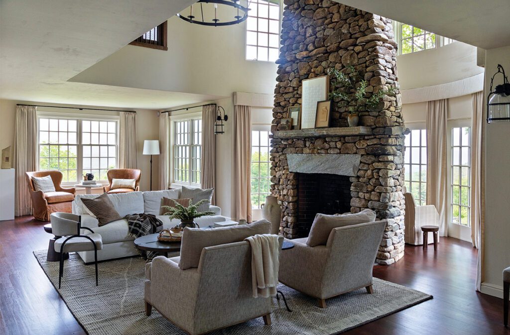 Spacious Vermont living room with a towering stone fireplace, neutral-toned furniture, large windows, and warm natural light creating a cozy and elegant atmosphere.