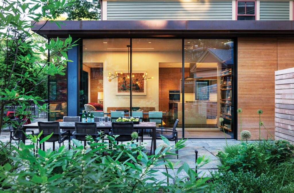 Modern outdoor dining area surrounded by lush greenery, featuring a sleek table with chairs, adjacent to a home with expansive glass walls that reveal a beautifully lit interior, including a dining space with contemporary decor and warm wooden accents.