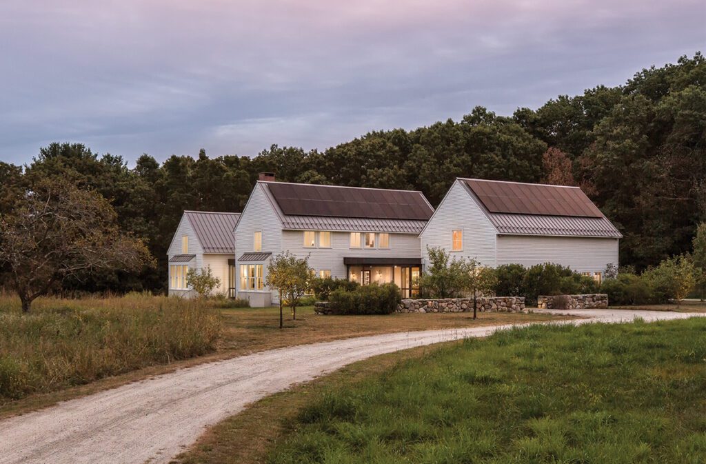 Side exterior of modern farmhouse with metal roof and illuminated windows at dusk by Elliott Architects.