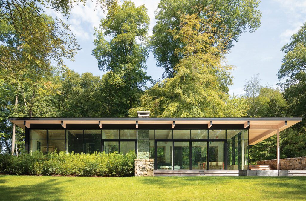 Modern glass house with flat roof, wood structure, and expansive lawn surrounded by trees.